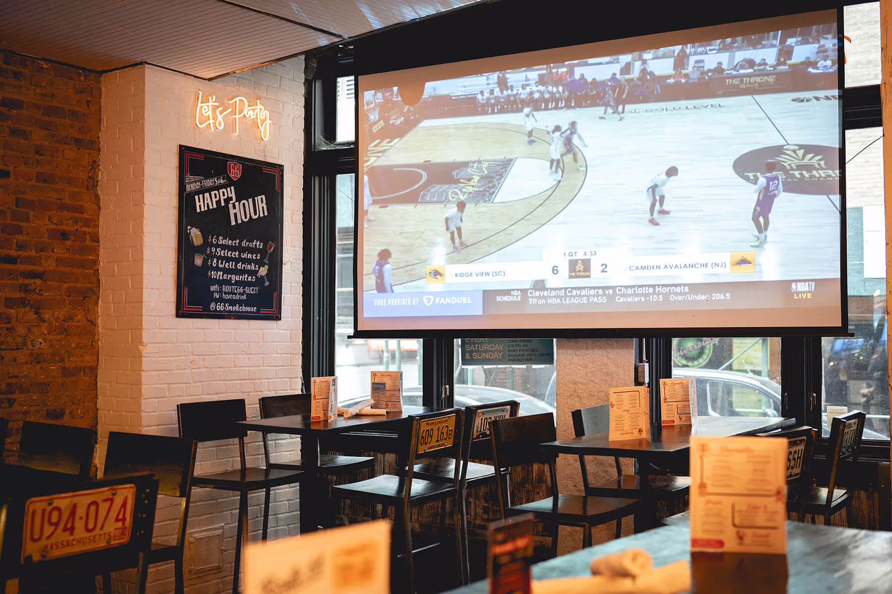 Interior of a sports bar with a large screen showing a basketball game and tables with menus and black chairs featuring vintage license plates.
