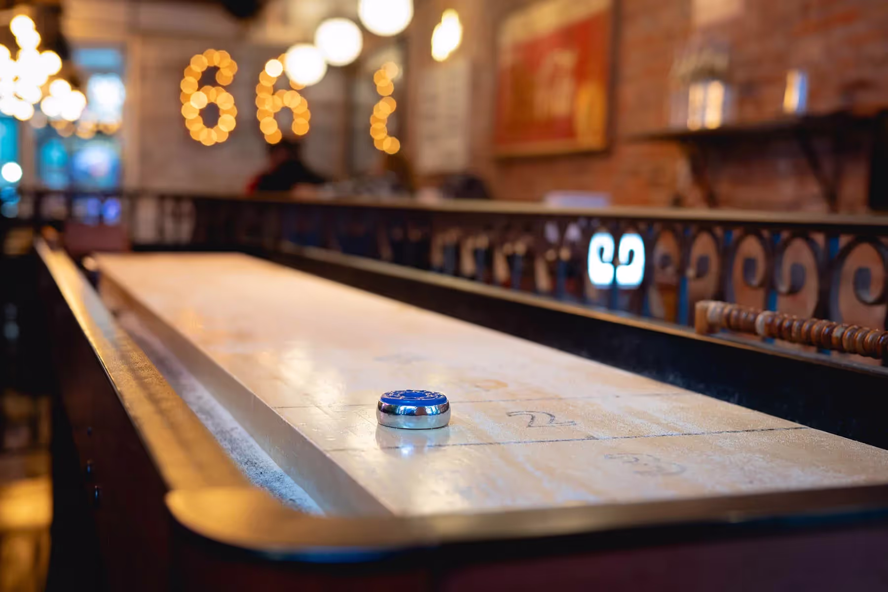 Close-up of a shuffleboard table with a blue puck on the playing surface in a softly lit bar interior.