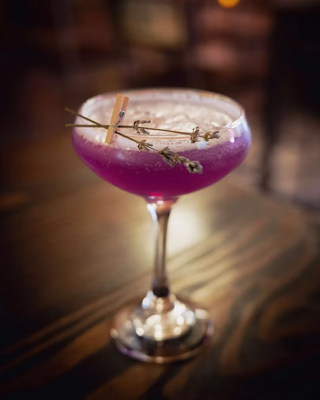 Purple cocktail in a coupe glass garnished with dried lavender sprigs clipped by a small wooden clothespin.