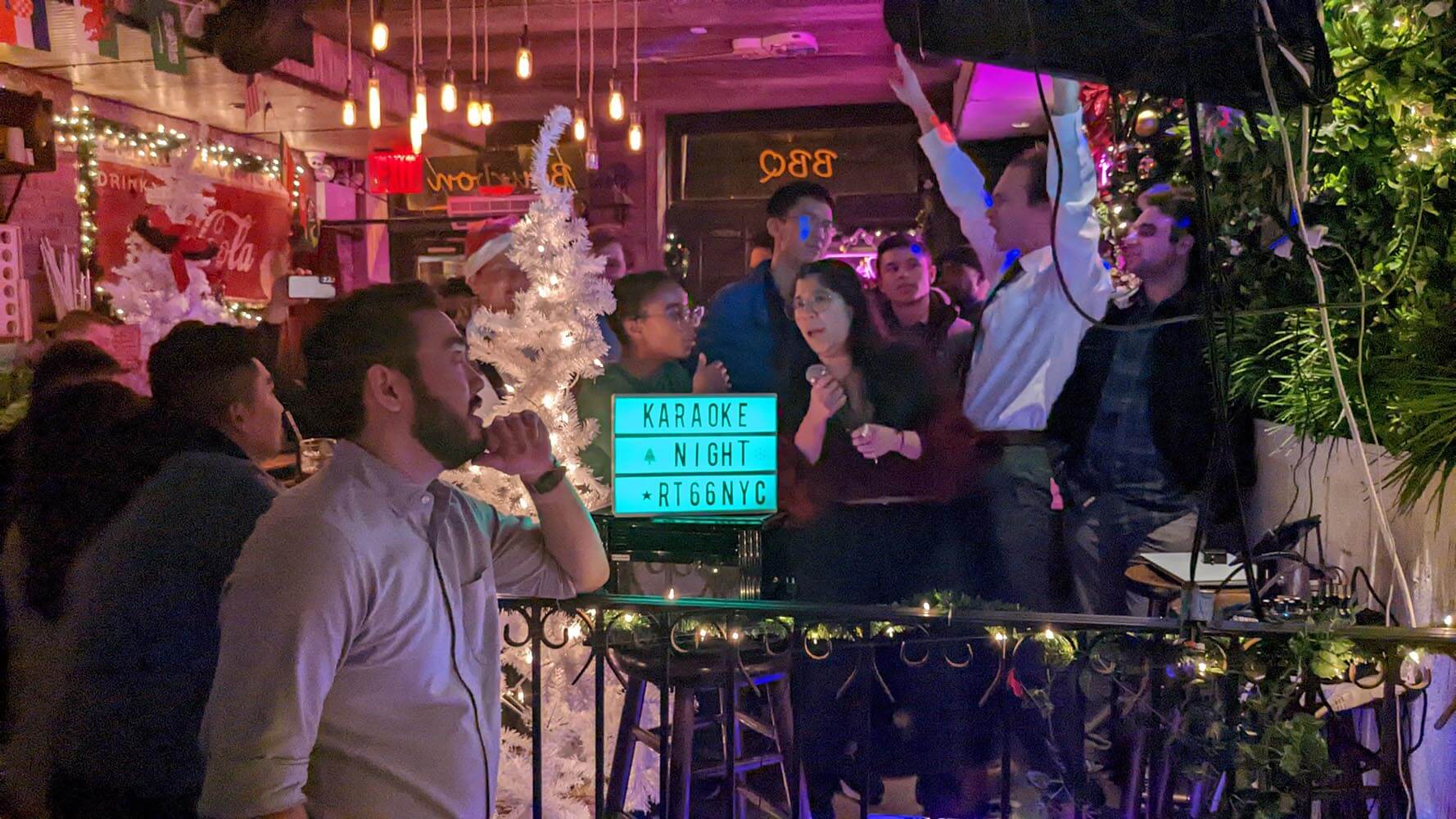 Group of people enjoying a karaoke night at a festive bar with Christmas decorations and a lighted sign displaying 'Karaoke Night RT66NYC'.