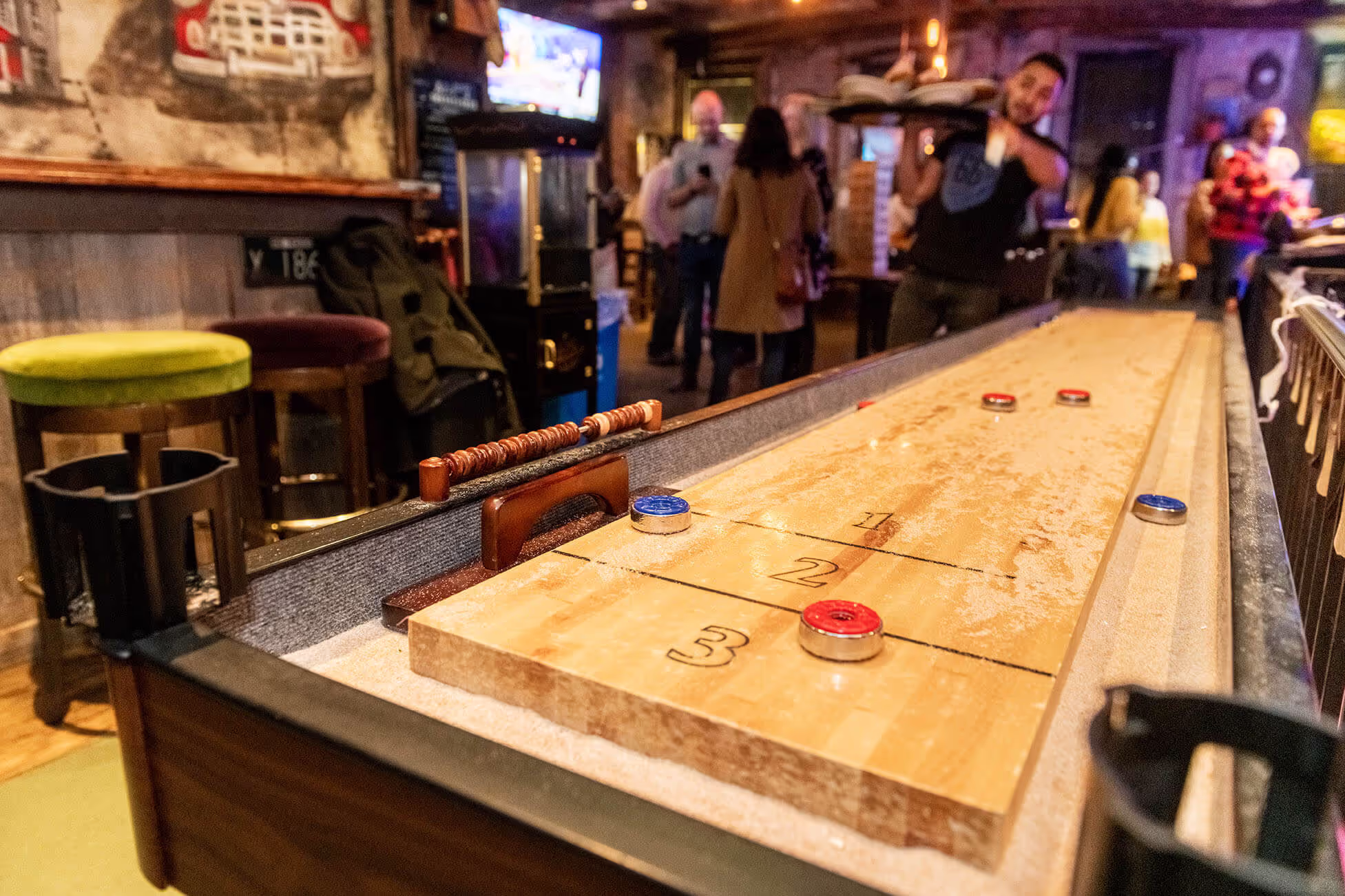 Shuffleboard table in a lively bar with colored pucks on the scoring area and people socializing in the background.