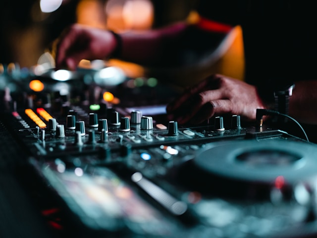 DJ hands adjusting knobs on a mixer in a dimly lit environment with blurred lights in the background.