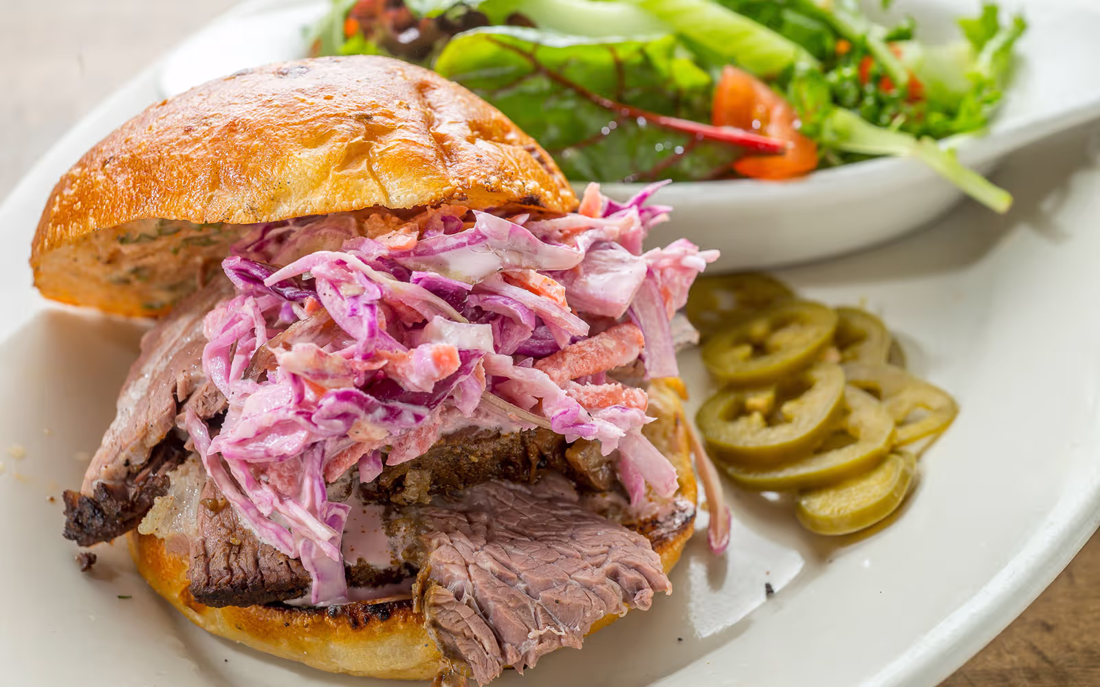 Beef sandwich with creamy purple coleslaw on a toasted bun, served with pickled jalapeño slices and a side salad.