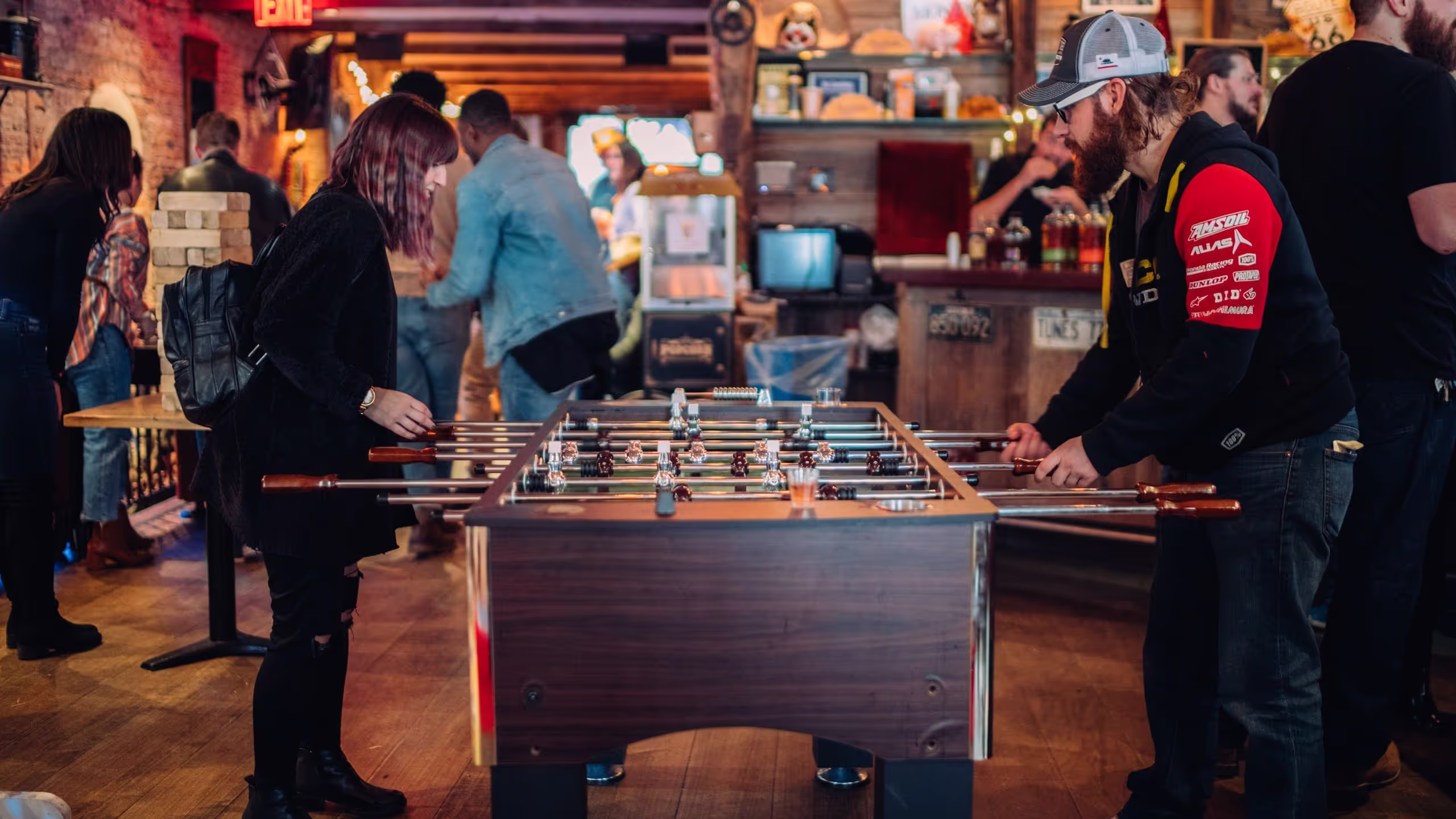 Two people playing foosball in a busy, warmly lit bar with other patrons in the background.