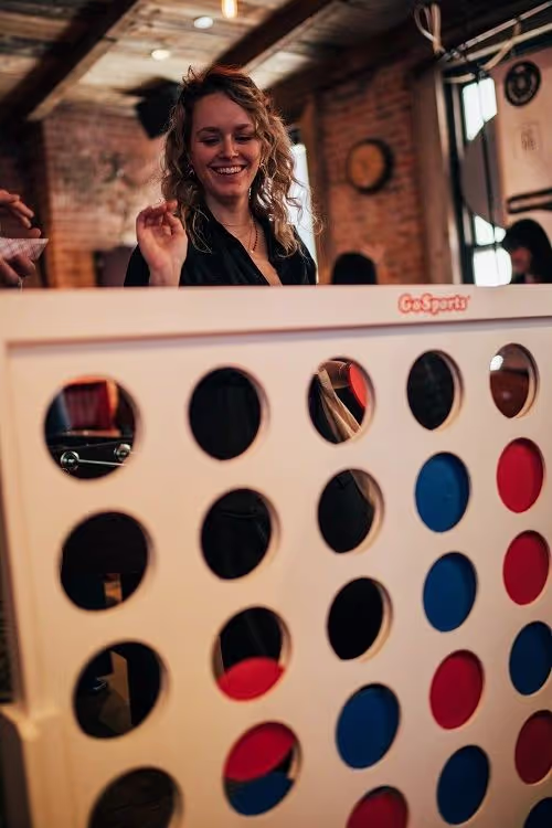 Smiling woman playing a large Connect Four game indoors with red and blue discs.