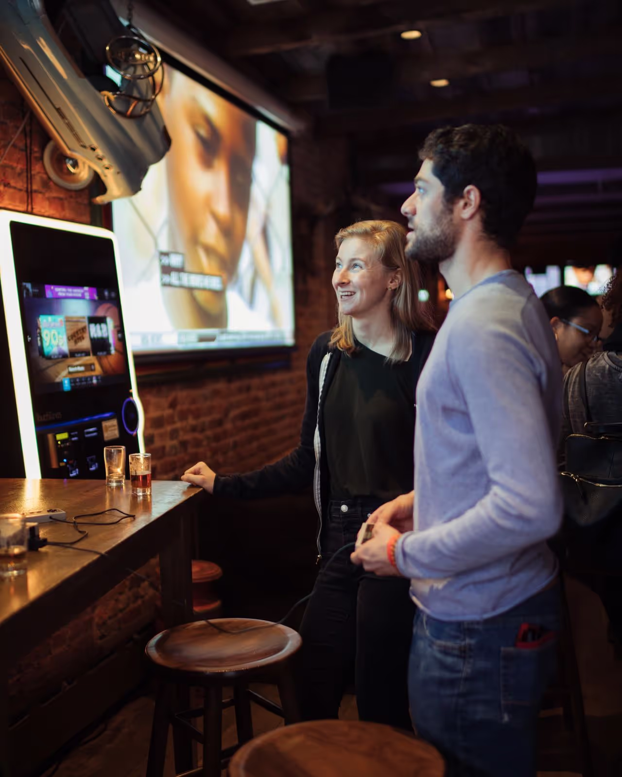 A man and woman smiling and playing video games together at a bar with stools and drinks on the counter.