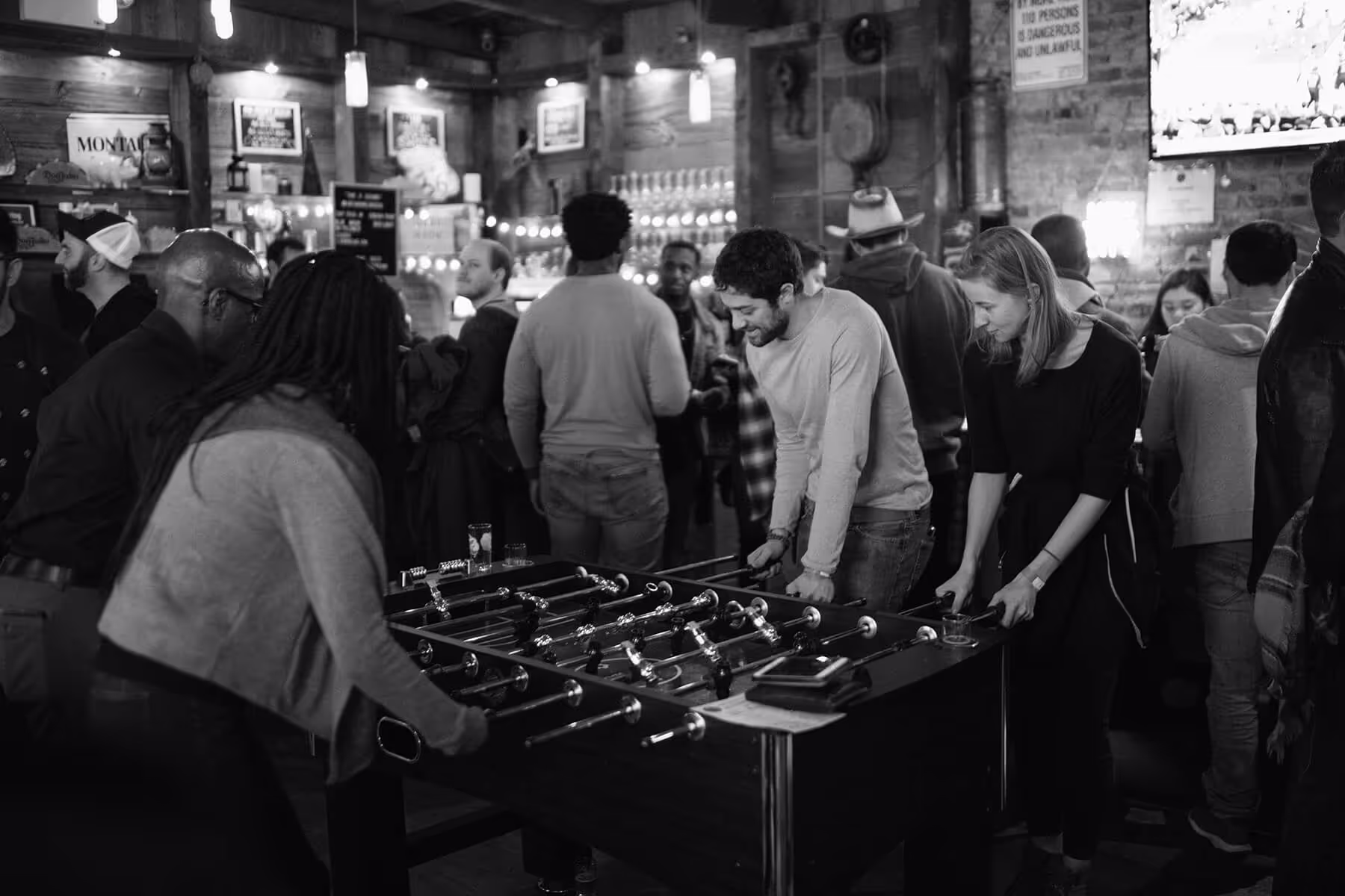 People playing foosball in a busy bar with others socializing in the background.
