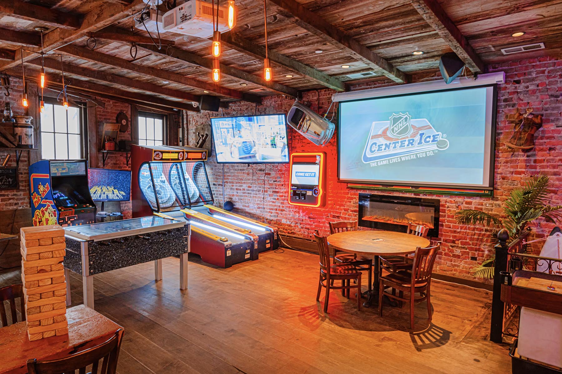 Cozy game room with exposed brick walls featuring arcade games, skee-ball machines, a large screen projector, round wooden table with chairs, and a Jenga block tower.
