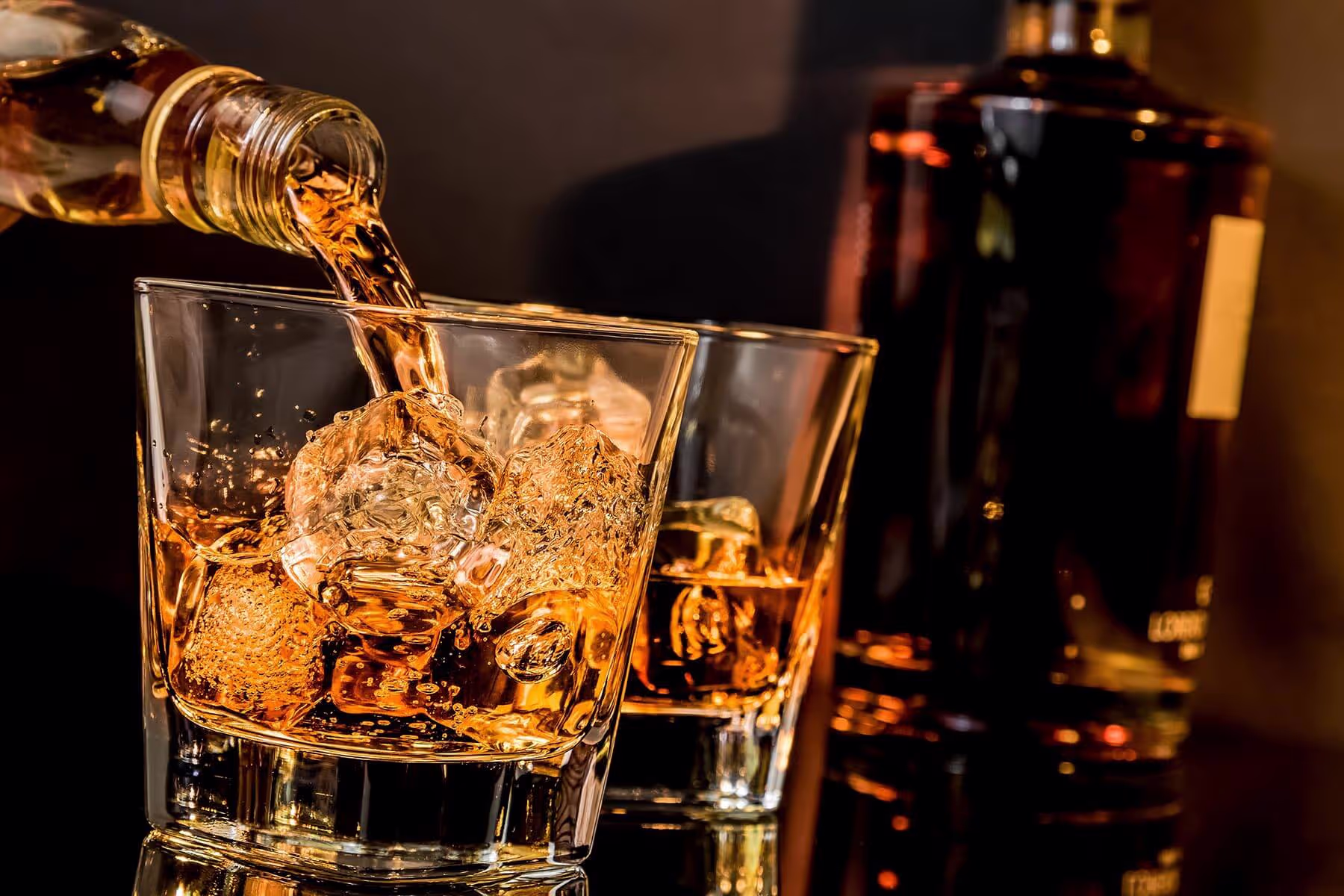 Whiskey being poured over ice cubes in a clear glass with another glass and bottle blurred in the background.